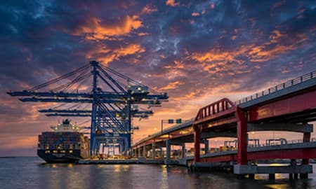 Grúas de una terminal de contenedores y un buque portacontenedores al atardecer, junto a un puente rojo